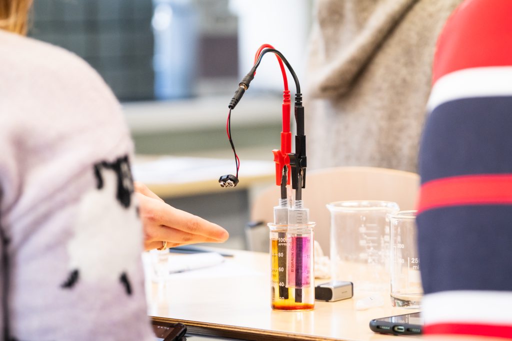 Lernende beobachten ein Elektrolyse-Experiment mit farbigen Lösungen im Klassenzimmer